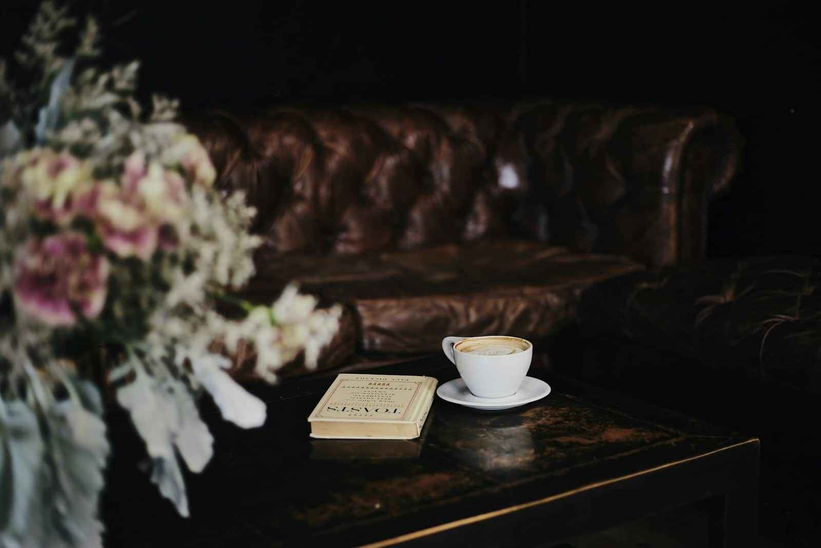 A cup of coffee and a closed book on a dark table in front of a leather sofa