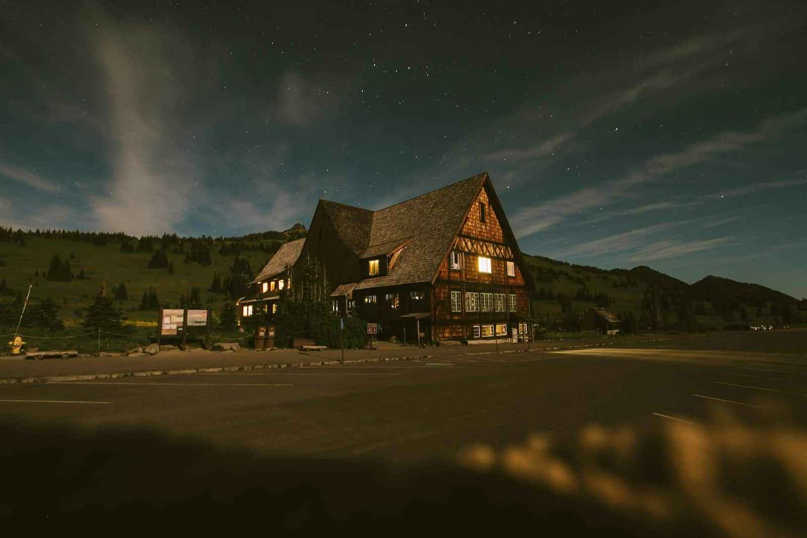 A timber house lit from the inside at dusk, standing in an open field with hills behind it