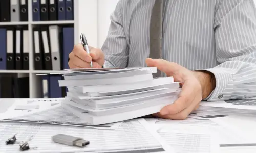  A man taking notes on top of thick paperwork with a USB flash drive and paperclip beside him.