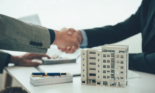 Two men exchanging a handshake over a miniature real estate building model.