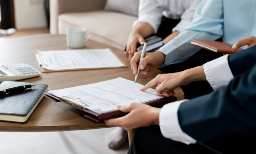 A group of three people in a meeting; one writing on paper, another pointing at documents, and the third listening attentively.