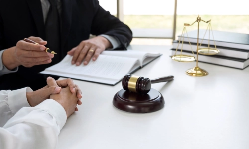An Albany injury lawyer discussing a case with a client while taking notes at a table with law books, a scale of justice, and a gavel hammer.